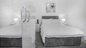 A modern hotel room with two beds, a gray headboard, white bedding, and a small bedside table with a lamp.