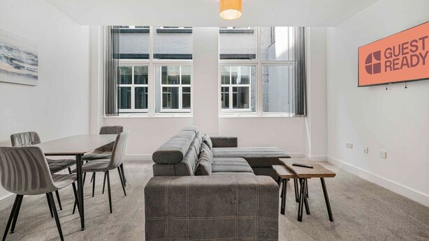 A modern hotel room with a large window, a sofa, a dining table, and a 'GUEST READY' sign.