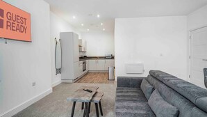 A modern, well-lit living area with a grey sofa, a small wooden table, and a kitchen in the background.