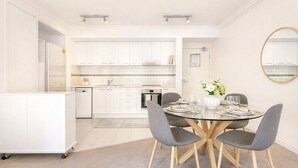 A modern kitchen with white cabinets, a dining area with a glass table, and grey chairs. - Walk to Elizabeth Quay | Pool & Parking (Perth)