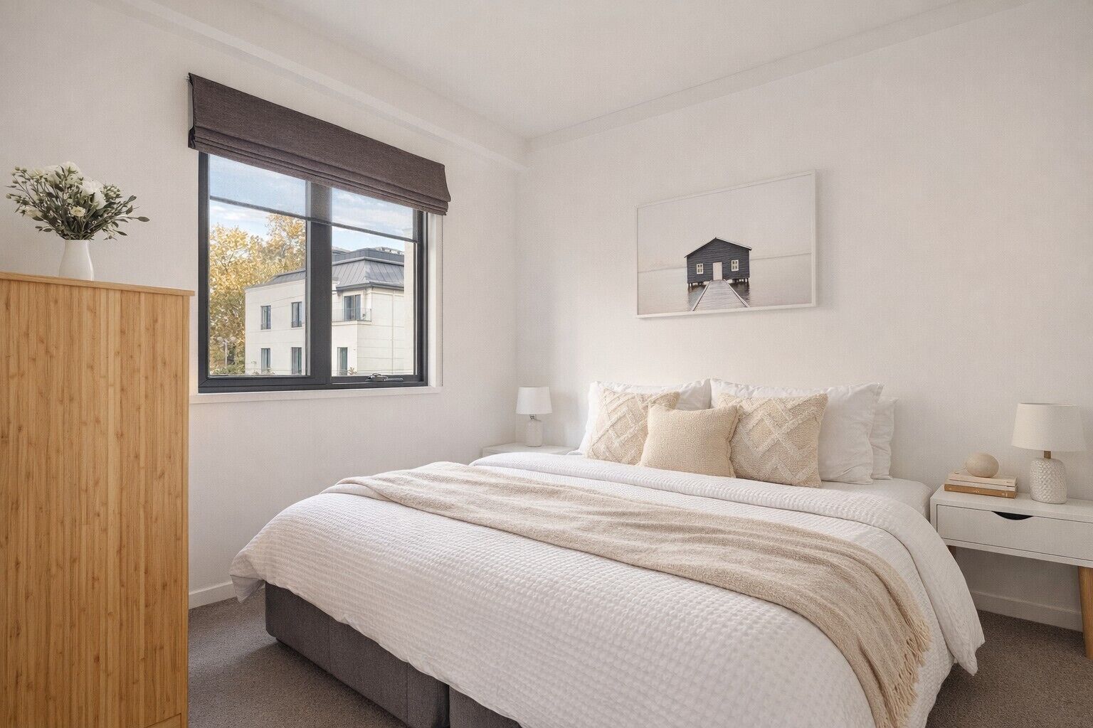 A bedroom with a large bed, a window with a view of buildings, a bedside table with a lamp, and a wall-mounted picture.