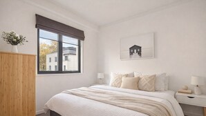 A bedroom with a large bed, a window with a view of buildings, a bedside table with a lamp, and a wall-mounted picture. - Tulizo  | Walk to Elizabeth Quay • Pool & Parking (Perth)