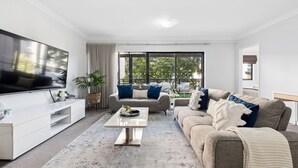 A modern living room with a flat-screen TV, a sofa, a coffee table, and a large window with a view of greenery. - Tulizo  | Walk to Elizabeth Quay • Pool & Parking (Perth)