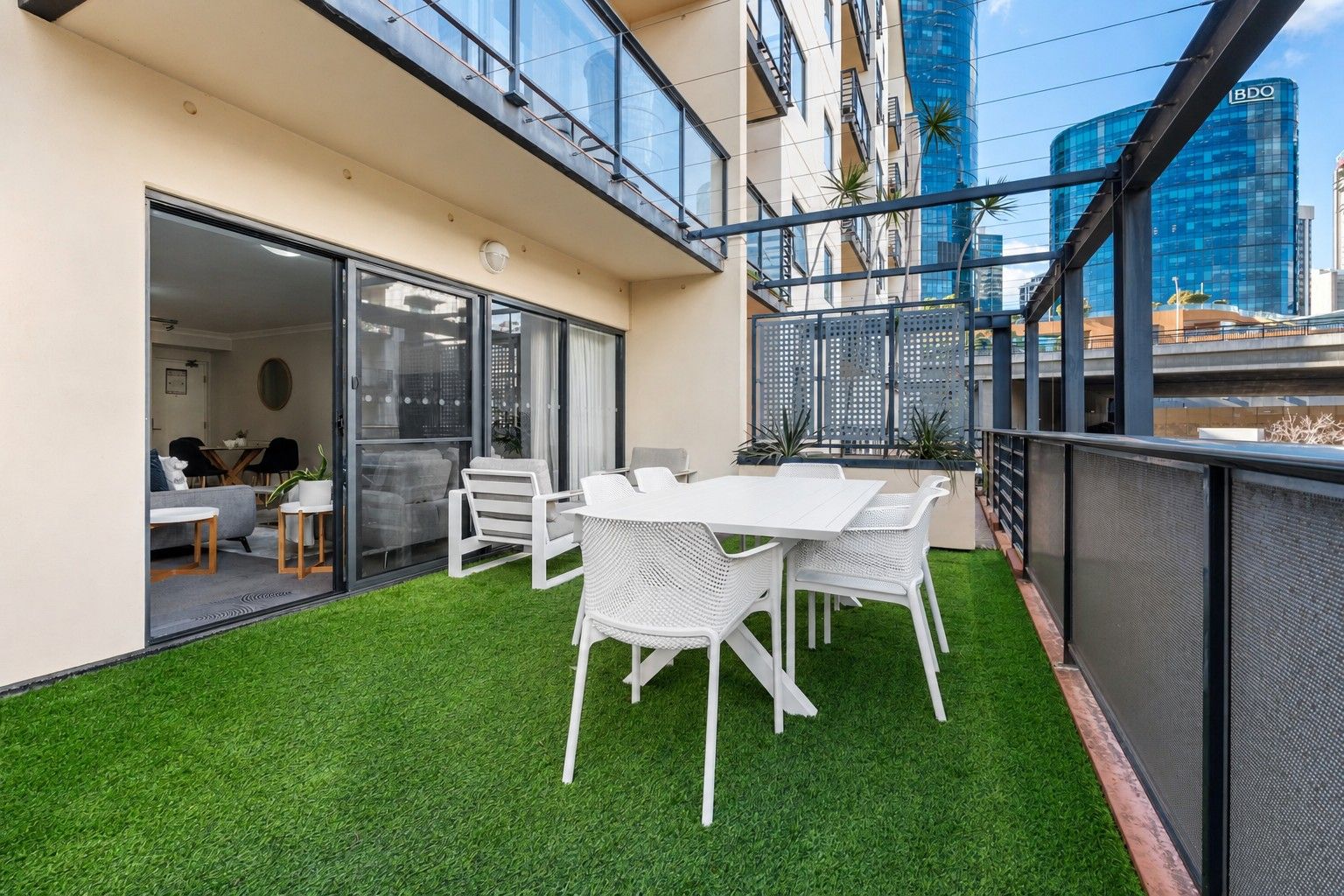 A modern outdoor patio with a white table and chairs, a glass door leading to an interior space, and a view of a cityscape with a tall building.