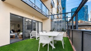 A modern outdoor patio with a white table and chairs, a glass door leading to an interior space, and a view of a cityscape with a tall building. - Tulizo  | Walk to Elizabeth Quay • Pool & Parking (Perth)