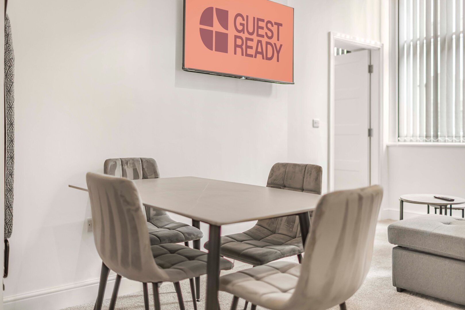 Una sala de conferències amb taula i quatre cadires, un cartell sobre la taula que diu “GUEST READY” i una finestra amb persianes a la dreta.