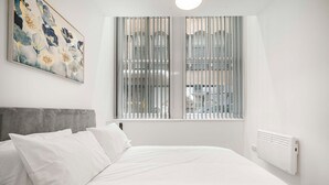 A bedroom with a bed, white bedding, a grey headboard, a window with blinds, a radiator, and a painting on the wall. - GuestReady – Modern comfort in Liverpool (Liverpool)