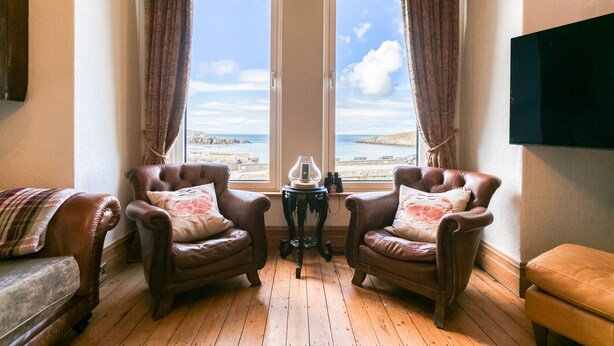 A room with two leather armchairs, a small table with a lamp, and a view of the beach through large windows.