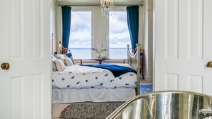 A bedroom with a bed, a small table with a vase, and a view of the sea through large windows.