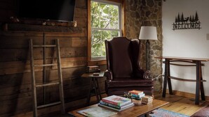A cozy room with a wooden interior, a stone wall, a large window, a leather armchair, a wooden table with books, a lamp, and a TV mounted on the wall. - The Serenity Cabin | Firepit | Wineries | Hiking (Marshall)