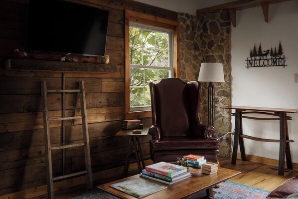 A cozy room with a wooden interior, a stone wall, a large window, a leather armchair, a wooden table with books, a lamp, and a TV mounted on the wall. - The Serenity Cabin | Firepit | Wineries | Hiking (Marshall)