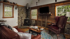 A cozy living room with a stone wall, a fireplace, a television, and a leather armchair. - The Serenity Cabin | Firepit | Wineries | Hiking (Marshall)