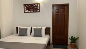 Cama bem arrumada com colcha branca e cinza, cabeceira de madeira e dois travesseiros. Uma peça decorativa está acima da cama, e uma porta de madeira fica à direita.