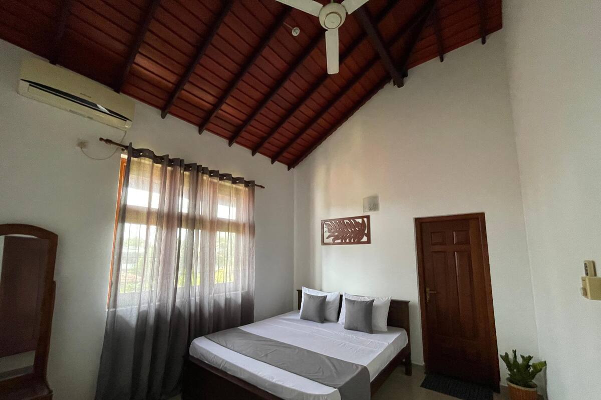 Une chambre à coucher comprenant un lit, des rideaux, un ventilateur de plafond, une porte et un élément décoratif fixé au mur.