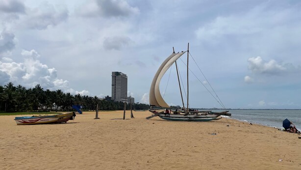 Uma praia com um veleiro, um edifício alto e palmeiras.