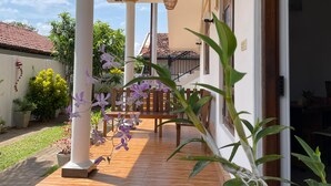 Varanda coberta com banco de madeira, plantas em vasos e vista para uma casa com telhado de cerâmica.