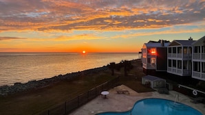 Une propriété située en bord de mer, dotée d’une piscine, d’une terrasse avec table et chaises, et offrant une vue sur le coucher de soleil sur l’océan.