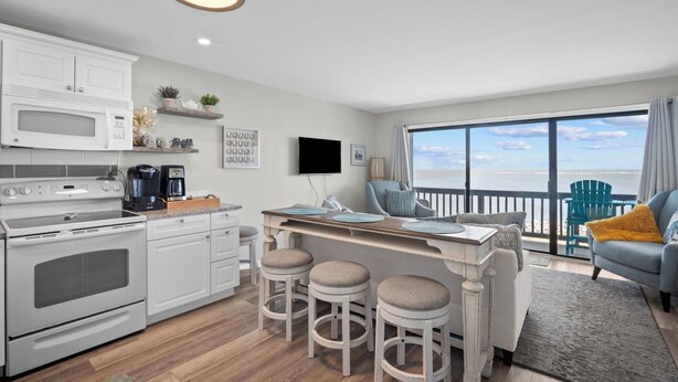 Une cuisine moderne avec des appareils électroménagers blancs, un coin bar avec des tabourets et un balcon offrant une vue sur l’océan.
