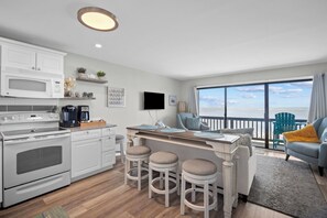 Une cuisine moderne avec des appareils électroménagers blancs, un coin bar avec des tabourets et un balcon offrant une vue sur l’océan.
