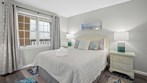 Une chambre à coucher avec un lit, deux tables de chevet, une fenêtre avec des rideaux et un tableau accroché au mur.