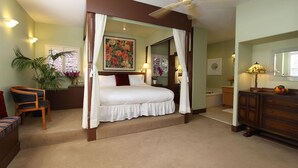 A four-poster bed with a canopy, a ceiling fan, a painting above the bed, and a desk with a lamp.