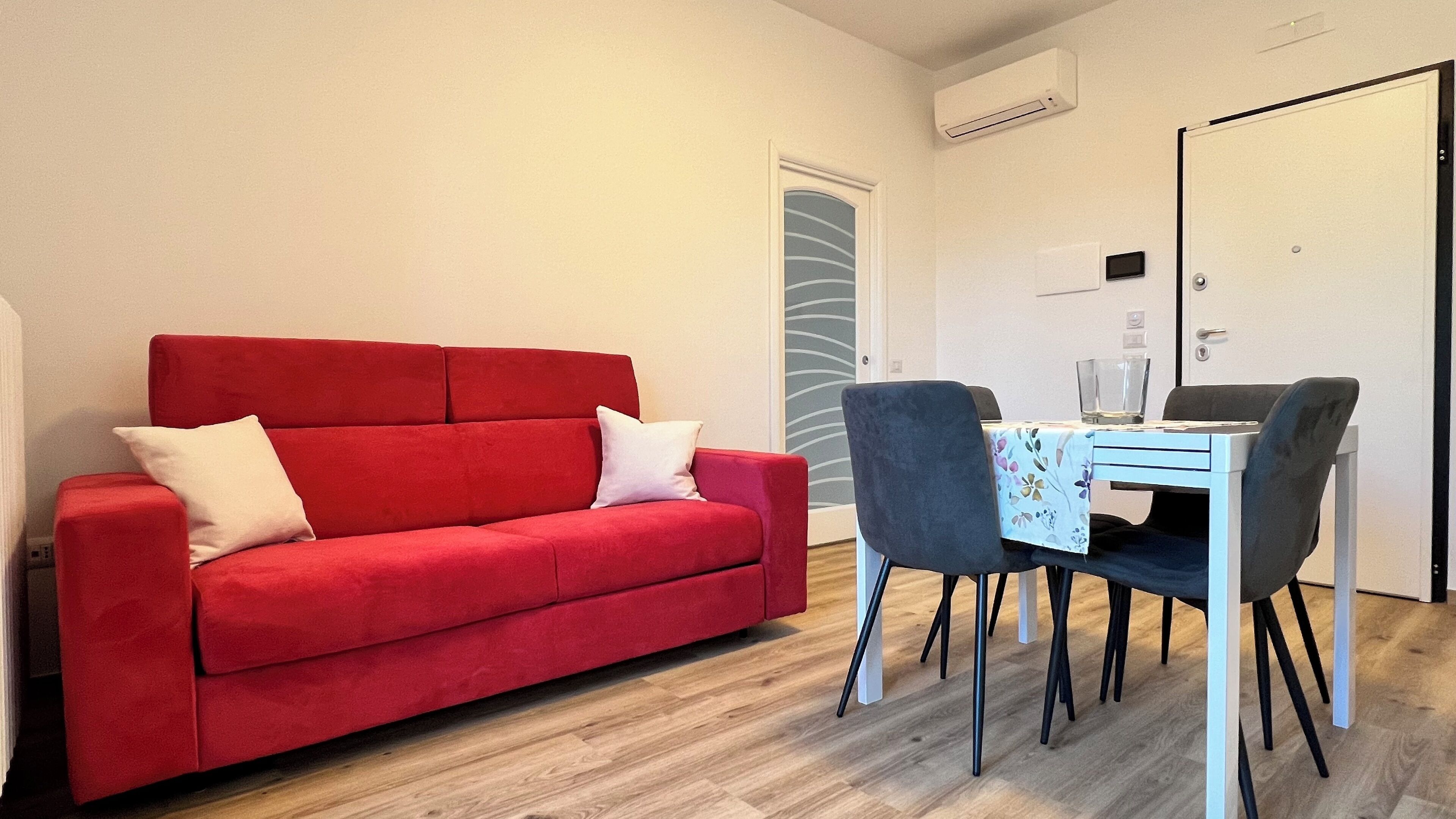 Ein rotes Sofa mit weißen Kissen, ein Esstisch mit Stühlen und eine Tür mit Griff.