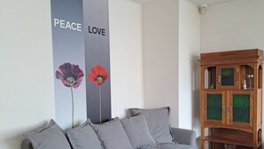 A grey sofa with grey cushions, a wooden cabinet with glass doors, and a wall with a vertical banner featuring flowers and the words "PEACE LOVE". - Cozy apt, 100m beach, AC, workstation, balcony, backyard, self check-in, yogamat (Jūrmala)
