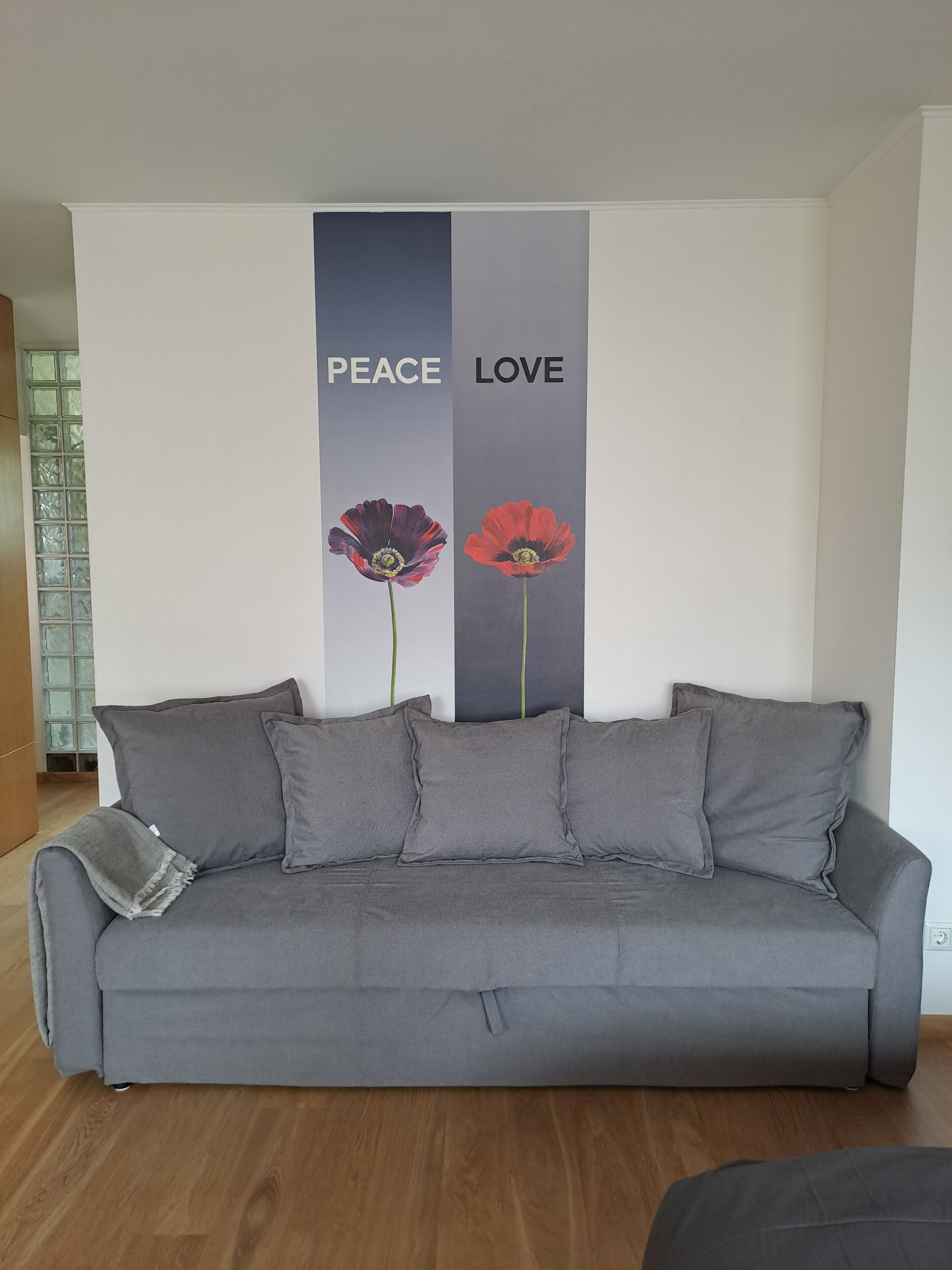 Un sofà gris amb tres coixins i una manta, col·locat davant d'una paret amb dos panells verticals que diuen "PEACE" i "LOVE" i que mostren imatges de flors vermelles i púrpures.