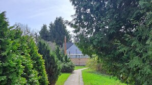 A paved pathway flanked by dense green foliage, leading to a building with a sloped roof. - Cozy apt, 100m beach, AC, workstation, balcony, backyard, self check-in, yogamat (Jūrmala)