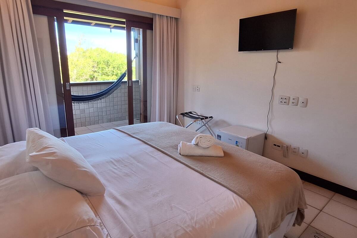 Una camera d'albergo con un letto, una televisione montata a parete, una finestra con tende e un tavolino con un asciugamano.