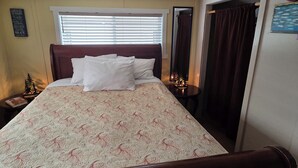 A neatly made bed with white linens and a floral-patterned duvet, a bedside table with a small lamp, a window with blinds, and a mirror on the wall. - Offut Lake!  Pet friendly cottage with lake access..  Bring your fishing rod! (Tenino)