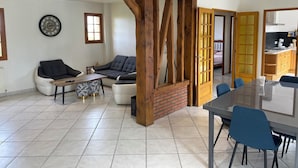 A spacious living area with a sofa, a coffee table, and a dining table with chairs. There is a wooden beam structure in the center, and a kitchen area is visible through an open door. - Ma belle Normande entre terre & mer - My beautiful Normandy between land and sea (Saint-Léonard)