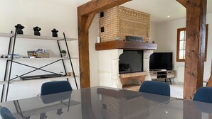 A modern living room with a glass-top dining table, blue chairs, a brick fireplace, and a TV. - Ma belle Normande entre terre & mer - My beautiful Normandy between land and sea (Saint-Léonard)