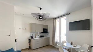 Un espacio habitable compacto con kitchenette, un comedor con sillas blancas, un sofá azul y un televisor montado en la pared.