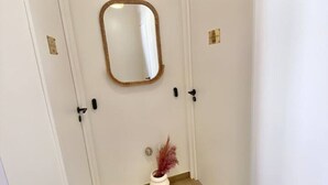 Un pasillo estrecho con paredes blancas, piso de madera, un espejo en la pared y un pequeño jarrón con plantas secas al final.
