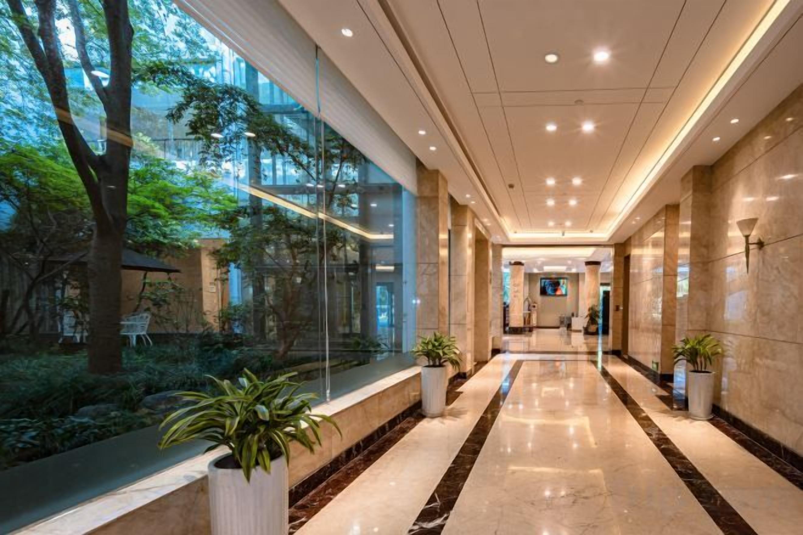 A modern hotel corridor with glass walls, potted plants, and a marble floor.