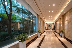 A modern hotel corridor with glass walls, potted plants, and a marble floor.