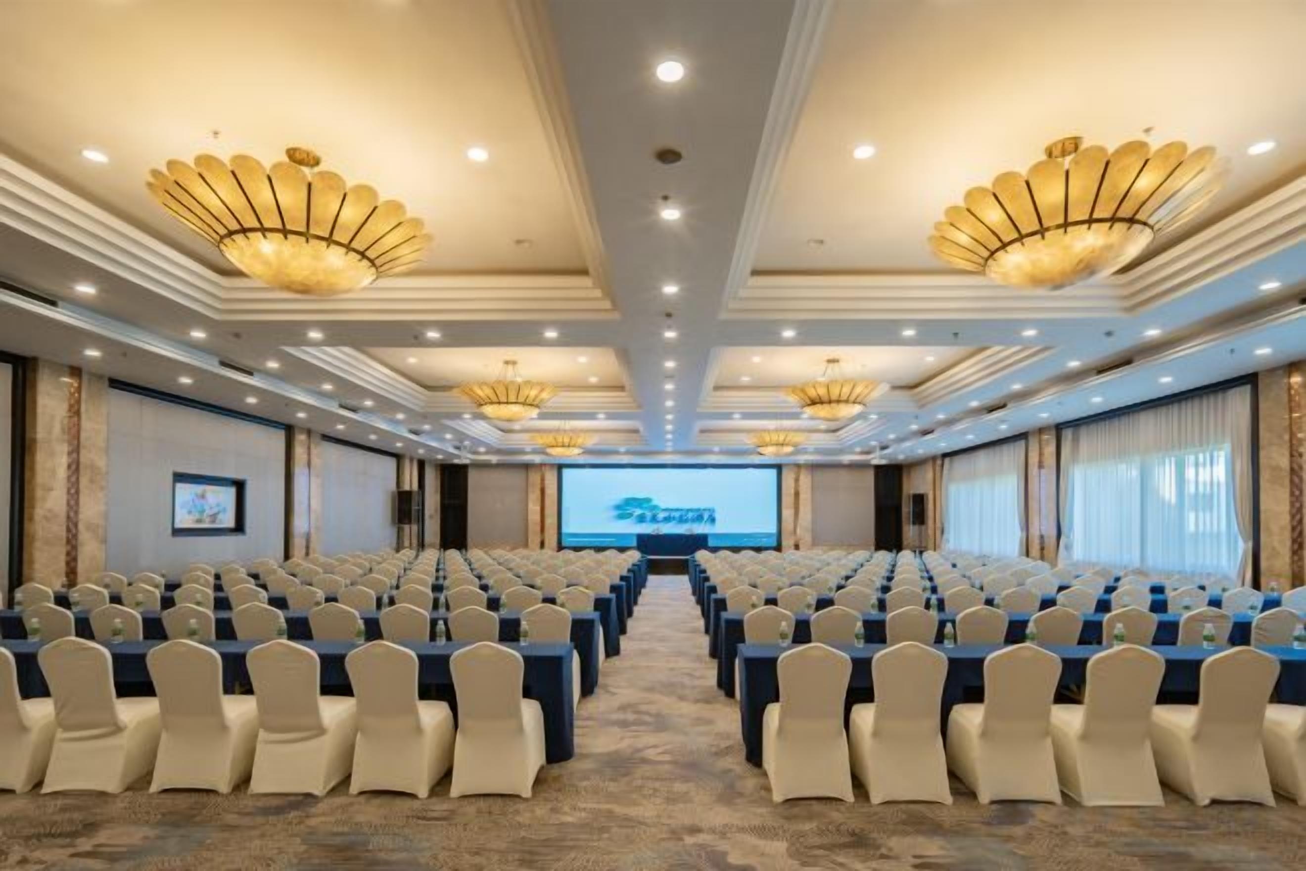 Un salón de conferencias con filas de sillas, una pantalla grande y candelabros.