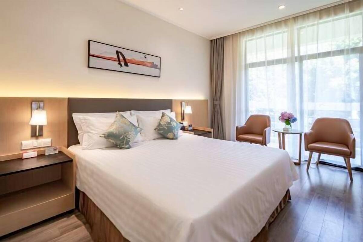A hotel room with a large bed, two bedside tables with lamps, a chair, a small table with a vase, and a window with curtains.