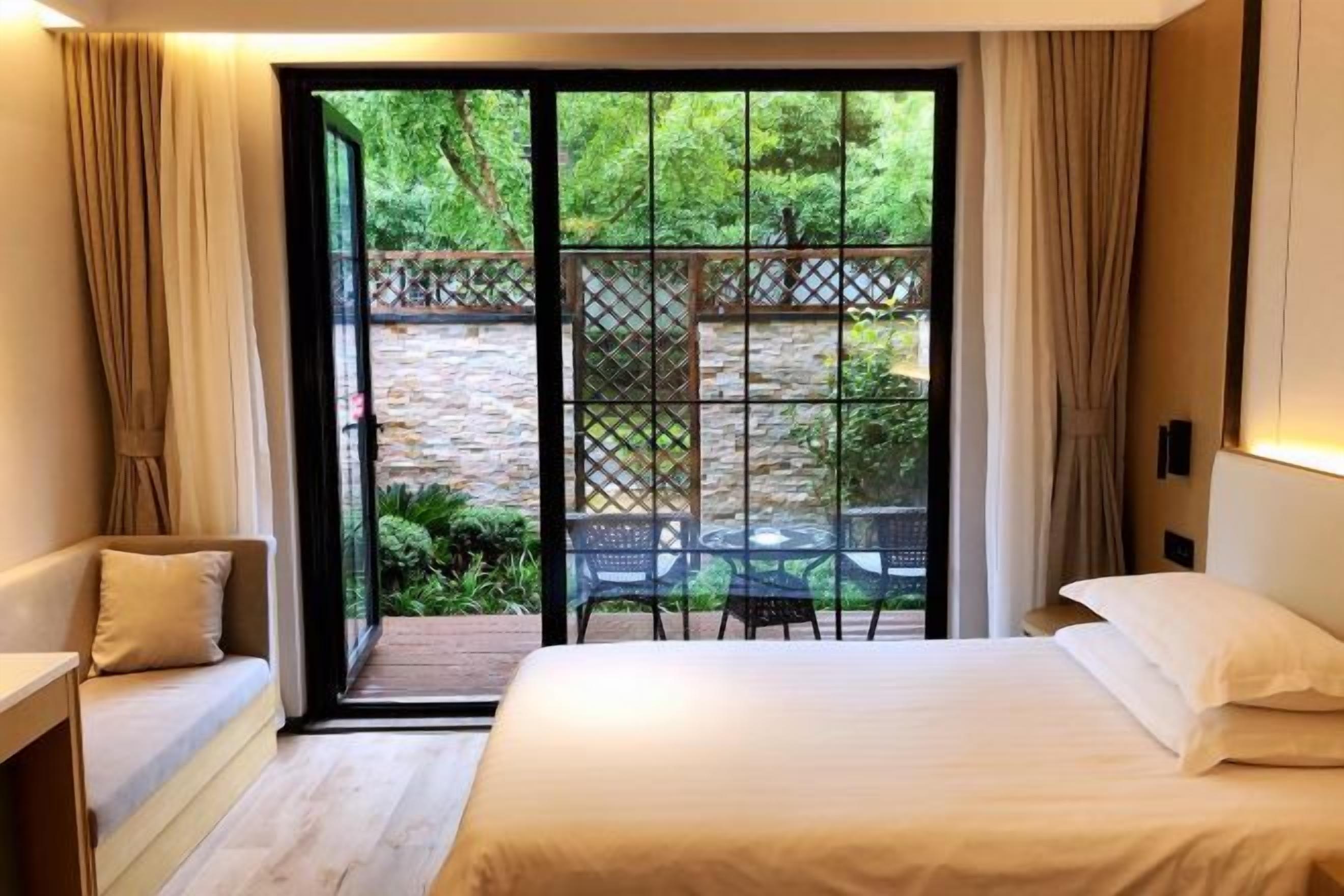 A bedroom with a large bed, a sofa, and a view of a garden through a glass door.