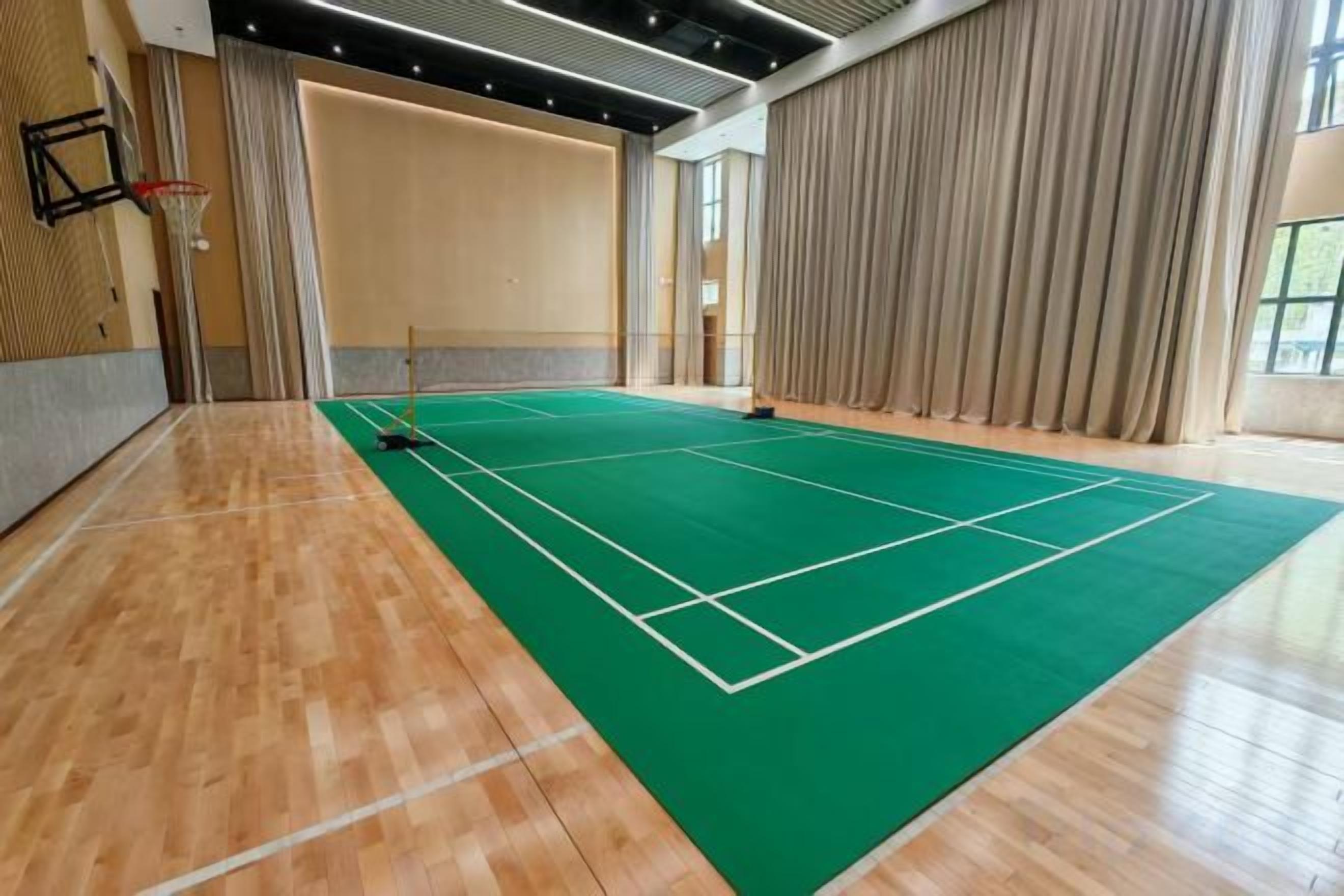 A tennis court with a wooden floor and a green playing surface, surrounded by curtains and a basketball hoop.