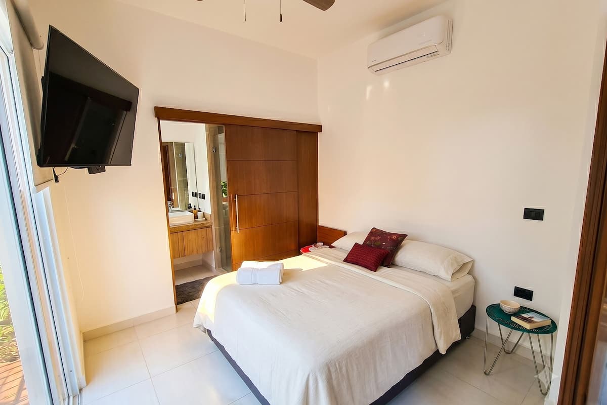 Quarto com cama, televisão, ar condicionado e uma pequena mesa com uma tigela e um livro.