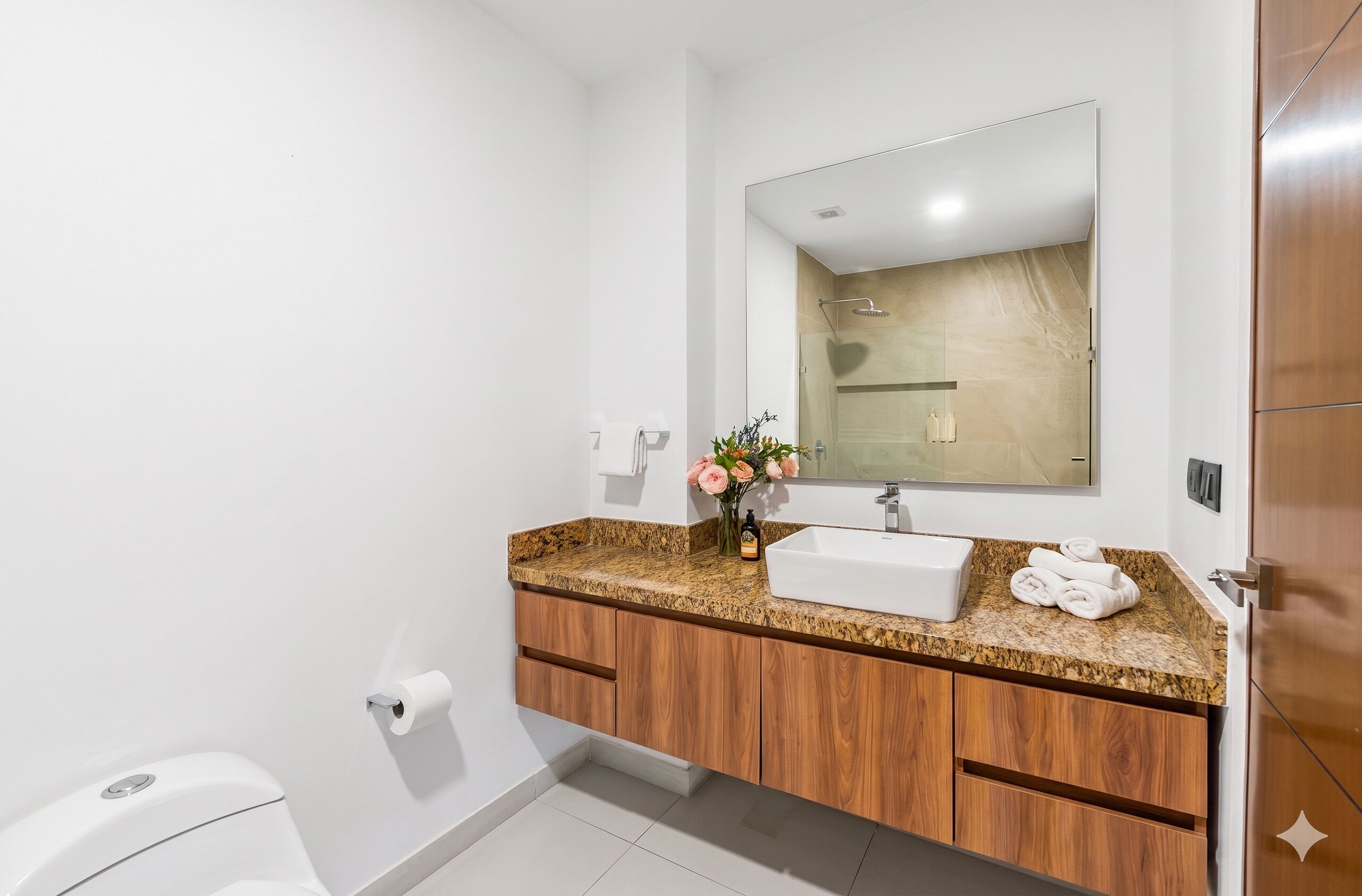 Un baño moderno con inodoro blanco, lavabo con encimera de granito, armarios de madera, un espejo y zona de ducha.