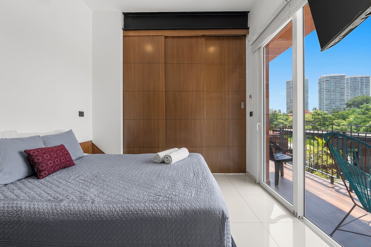 A modern bedroom with a large bed, a wooden wardrobe, and a balcony with a view of the city.