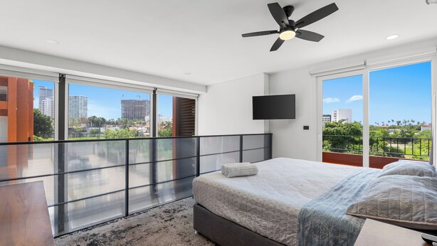 A modern bedroom with a large bed, a ceiling fan, a TV mounted on the wall, and a balcony with a view of the city. - Zoho Skies by Monyxbnb (Puerto Vallarta)