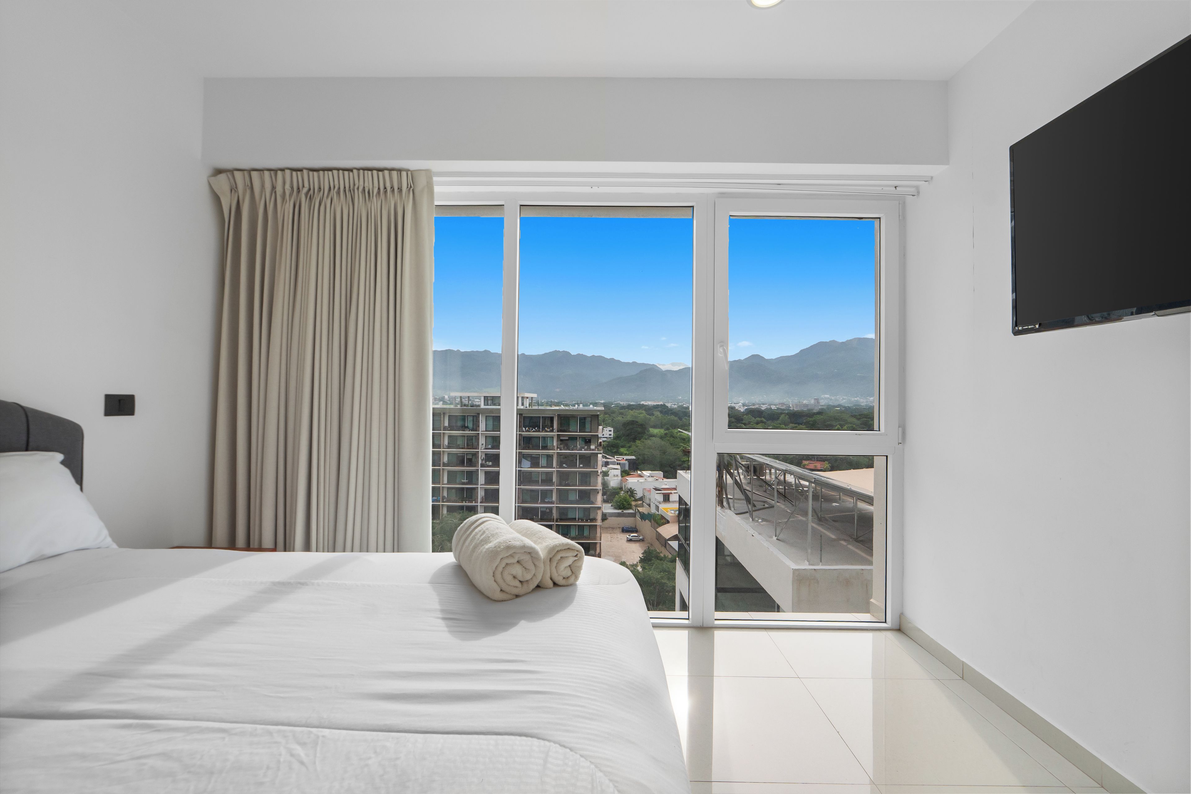 Habitación de hotel con una cama grande, vista a un paisaje urbano y un balcón con montañas al fondo.