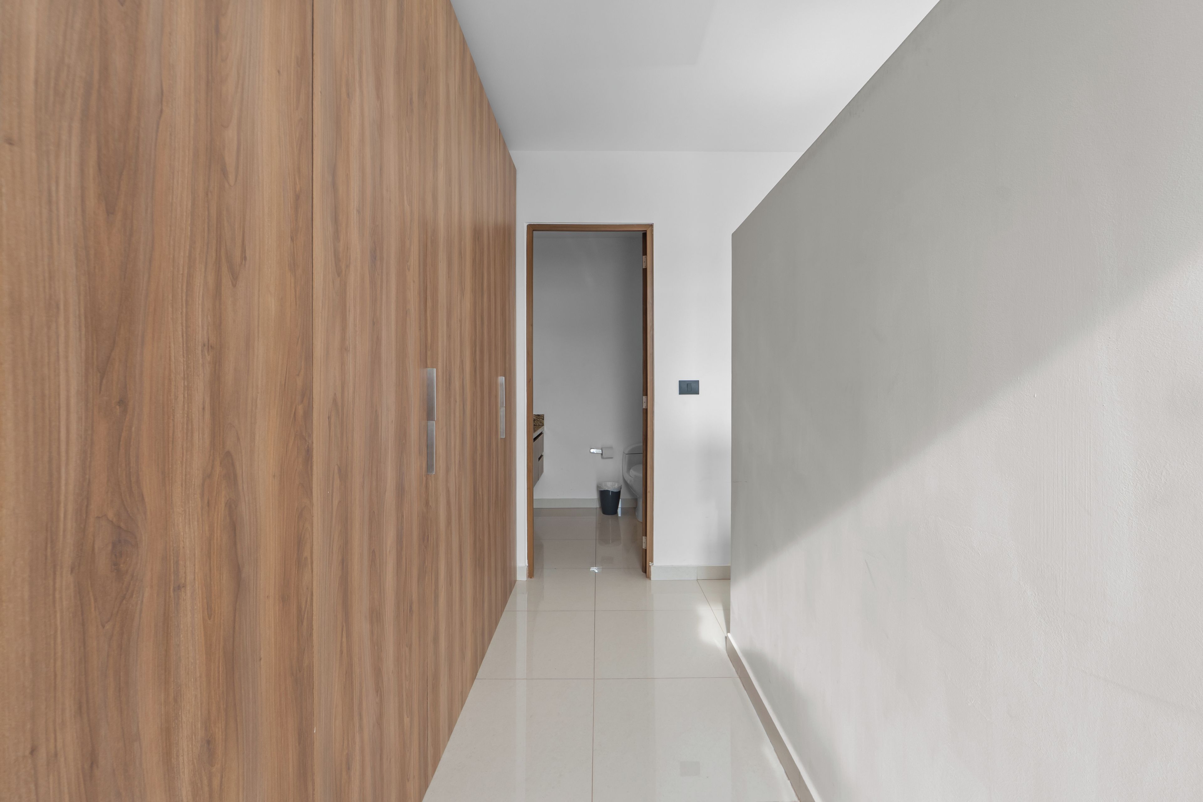 A hallway with a wooden wardrobe, a white wall, and a door leading to a bathroom.