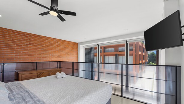 A modern bedroom with a large bed, a ceiling fan, a brick wall, and a balcony with a view of buildings. - Zoho Skies by Monyxbnb (Puerto Vallarta)