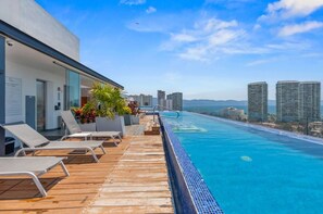 Una piscina infinita en la azotea con sillones de descanso, un muelle de madera y vistas a rascacielos.