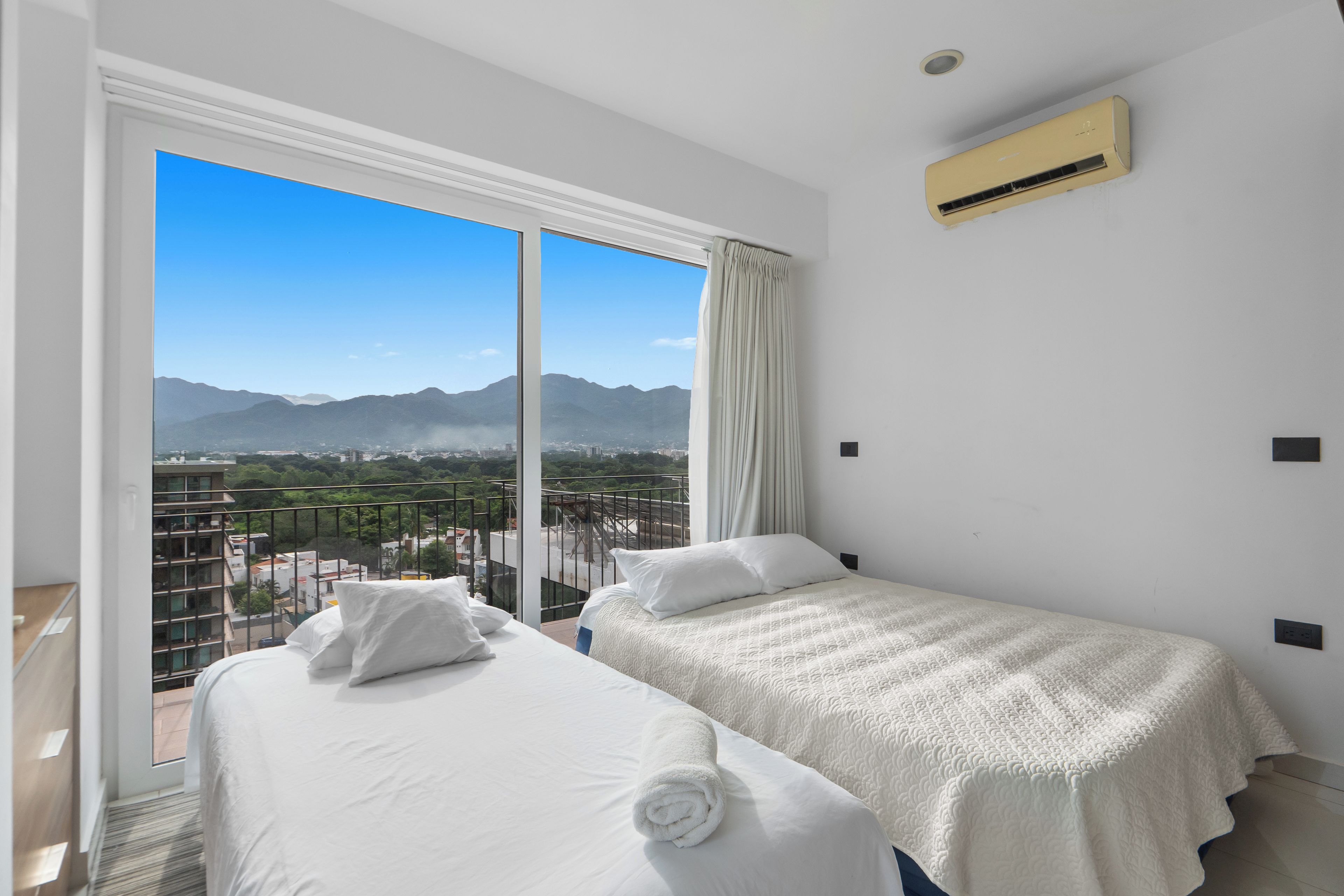 A hotel room with two beds, a large window with a view of mountains, and an air conditioning unit on the wall.